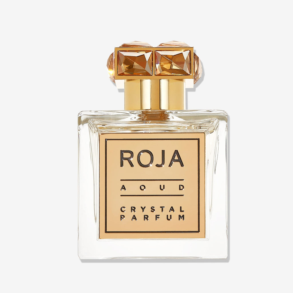 Aoud CRYSTAL™️ by Roja Parfums is a clear glass perfume bottle with a gold cap adorned by two gem-like decorations and a gold-bordered label, capturing luxury with opulent Saffron & Rose Oud notes.