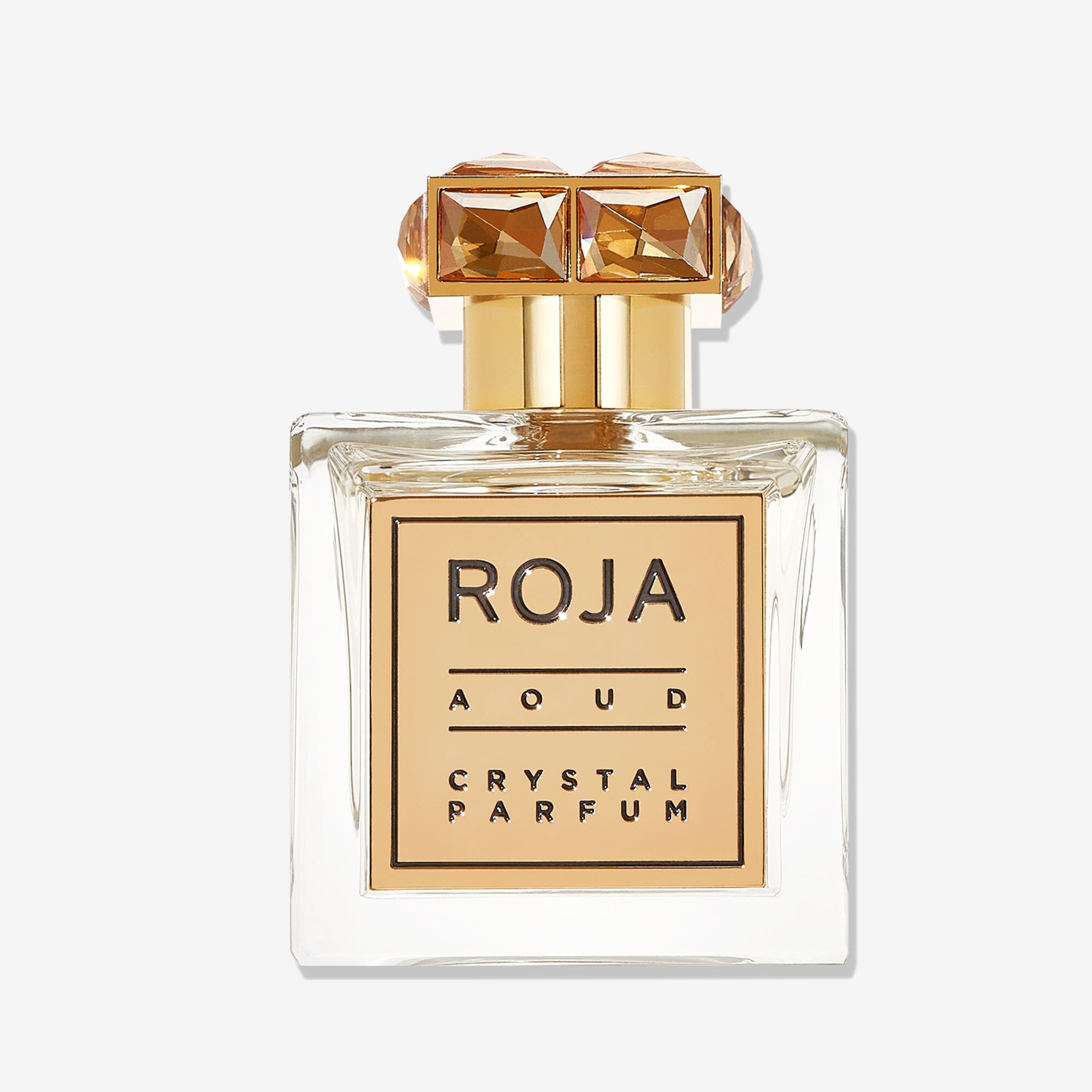 Aoud CRYSTAL™️ by Roja Parfums is a clear glass perfume bottle with a gold cap adorned by two gem-like decorations and a gold-bordered label, capturing luxury with opulent Saffron & Rose Oud notes.